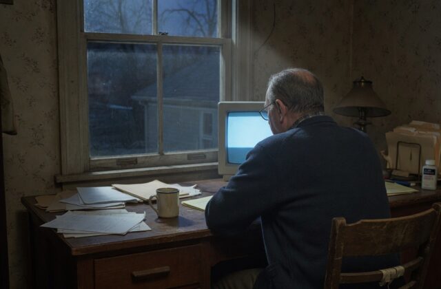 A man working at a computer at a desk by a window in a dim, old-fashioned room
